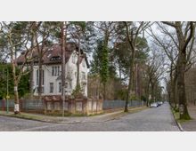 Straßenszene in einem Wohngebiet mit Kopfsteinpflaster, großen Bäumen und geparkten Autos. Auf der linken Seite befindet sich ein großes, mehrstöckiges Haus mit weißer Fassade und rotem Dach, das von einem Backsteinzaun umgeben ist.