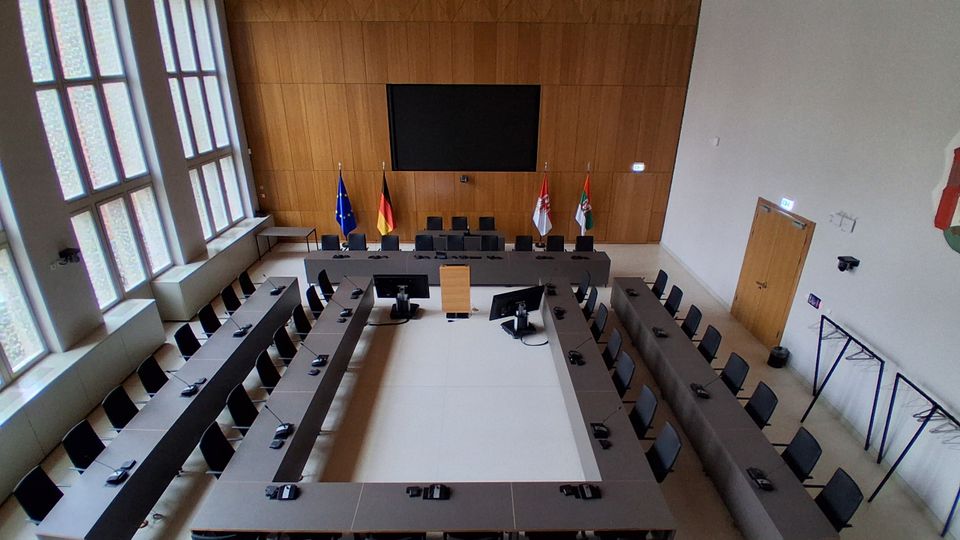 Moderner Sitzungssaal mit U-förmig angeordneten Tischen und zahlreichen Stühlen, ausgestattet mit Mikrofonen und Monitoren. Vor einer holzverkleideten Wand hängen ein großer Bildschirm und mehrere Flaggen; links sorgen hohe Fensterfronten für Tageslicht.
