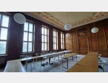 Innenansicht eines historischen Besprechungsraums mit holzvertäfelten Wänden, hohen Fenstern mit dekorativen Gittern und einer verzierten Decke. Im Raum stehen Tische und weiße Kunststoffstühle; von der Decke hängen runde, weiße Leuchten.