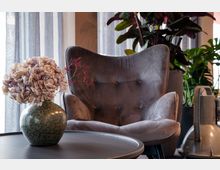 Ein gemütlicher Innenraum mit einem grauen Sessel im Hintergrund, dekoriert mit großen Zimmerpflanzen. Im Vordergrund steht ein runder Tisch mit einer Vase, die getrocknete Blumen und Zweige enthält.