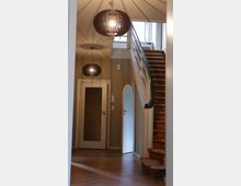 Ein Eingangsbereich mit Holztreppe, die nach oben führt, einer offenen Tür und zwei modernen, dunklen Hängelampen an der Decke. Der Boden besteht aus Holz, und rechts neben der Treppe befindet sich ein großes Fenster, das natürliches Licht hereinlässt.