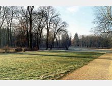 Weitläufige Parklandschaft mit einer gepflegten Rasenfläche im Vordergrund, einem Sandweg am rechten Bildrand und einer Reihe von laublosen Bäumen im Hintergrund, die an einen lichten Wald grenzen. Eine niedrige steinerne Balustrade befindet sich links im Bild.