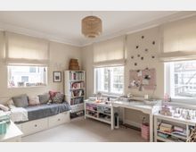 Helles Jugendzimmer mit beigen Wänden und großen Fenstern mit hellen Rollos. Zu sehen sind eine Tagesbettcouch, ein Bücherregal, ein Schreibtisch mit Dekoration, sowie persönliche Gegenstände und Polaroid-Fotos an der Wand.