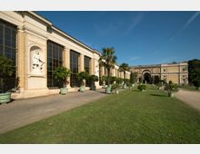 Historisches Gewächshaus mit großen Glasfenstern und verzierten Backsteinmauern, flankiert von Skulpturen in Nischen; vor dem Gebäude stehen geometrisch angeordnete Kübelpflanzen und Palmen auf einer gepflegten Rasenfläche. Im Hintergrund ist ein Torbogen zwischen zwei Gebäudeteilen zu sehen.