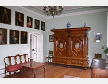 Historischer Raum mit holzgetäfeltem Boden, einem großen verzierten Kleiderschrank aus Holz, einer antiken Sitzbank mit roter Polsterung und einem quadratischen Holztisch. Porträts historischer Persönlichkeiten hängen an den hellgrünen Wänden, und eine dekorative Deckenleuchte hängt zentral im Raum.