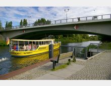 Eine gelbe, überdachte Fähre mit Passagieren fährt unter einer modernen Betonbrücke auf einem Fluss hindurch. Im Vordergrund befinden sich ein gepflasterter Gehweg, eine Holzbank und ein Geländer; im Hintergrund ist grüne Ufervegetation zu sehen.