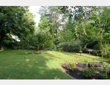 Ein Garten mit einer gepflegten Rasenfläche, einzelnen Obstbäumen und einem Blumenbeet im Vordergrund. Im Hintergrund sind dichte Sträucher und hohe Bäume zu sehen, mit einem Wasserstrahl aus einem Rasensprenger auf der Wiese.