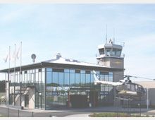 Modernes Flughafengebäude mit einer Glasfassade und einem angebauten Kontrollturm. Im Vordergrund steht ein ausgestellter Hubschrauber auf einem Podest, umgeben von einer gepflasterten Fläche und Fahnenmasten.