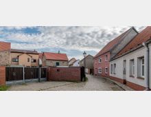 Eine schmale Kopfsteinpflasterstraße mit kleinen Wohnhäusern und roten Ziegeldächern auf beiden Seiten. Im Vordergrund befindet sich ein Tor mit Ziegel- und Holzelementen, während eine Laterne und eine durchwachsene Wolkendecke im Hintergrund sichtbar sind.