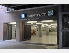 Eingang zur U- und S-Bahn-Station „Bundesplatz“ in Berlin, mit grauer Steinfassade, großen schwarzen Toren und Wegweiser „Richtung Osloer Straße“. Im Inneren sind beleuchtete Flure mit weißen Fliesen und Werbeplakaten an den Wänden erkennbar.