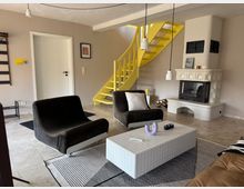 Wohnzimmer mit modernem Interieur, bestehend aus zwei braunen Sesseln, einem weißen Fliesentisch und einer weißen Kachelofen-Kaminanlage. Im Hintergrund befindet sich eine gelb gestrichene Wendeltreppe und eine Tür in einer cremefarbenen Wand.