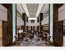 Hotel-Lobby mit modernem, luxuriösem Design: Große Marmorsäulen mit dunkler Maserung, helle Deckenbeleuchtung, Sitzbereiche mit Sesseln, Tischen und Lampen auf einem Teppich mit abstraktem Muster; im Hintergrund eine gläserne Balustrade und ein dekorativer Uhrenbereich.