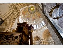 Innenansicht einer barocken Kuppelkirche mit reich verzierten Fresken, filigranen Stuckarbeiten und großen Fenstern, die Tageslicht einlassen. Im Vordergrund steht eine hölzerne Kanzel mit geschnitzten Details, flankiert von dekorativen Säulen.