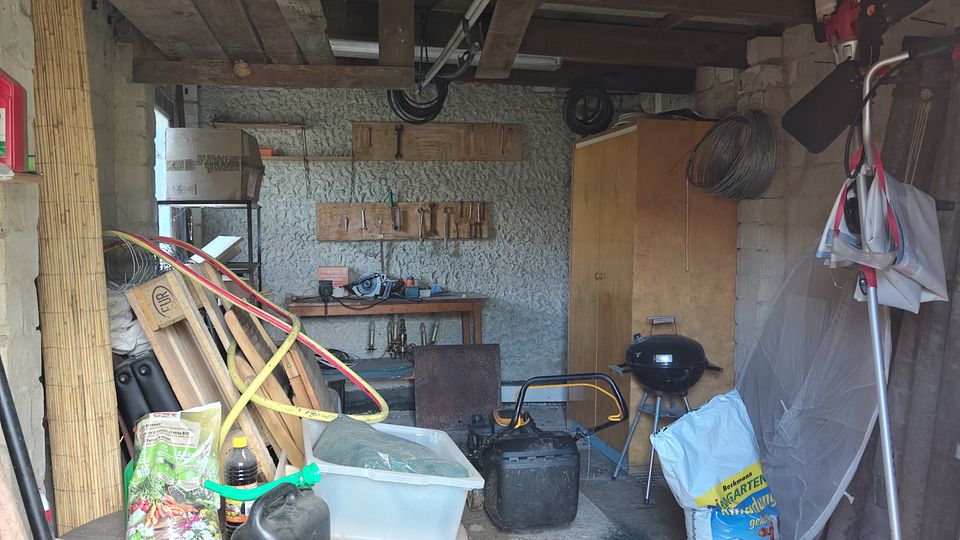 Innenansicht einer einfachen Garage bzw. eines Schuppens mit sichtbaren Holzbalken an der Decke, Betonwänden und einem gemusterten Bodenbelag. Der Raum ist mit gelagerten Gegenständen wie Tisch, Werkzeugbank, Schrank, Säcken, Kanistern, Brettern und einem Kugelgrill vollgestellt.