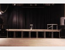 Bühne in einem Veranstaltungsraum mit schwarzem Vorhang als Hintergrund, mehreren Scheinwerfern an der Decke und einem einzelnen Barhocker auf der Bühne. Links und rechts stehen Lautsprecher, der Boden ist schlicht und unbedeckt.