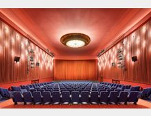 Ein Kino mit blauen Sitzreihen, roten Wänden und einem geschlossenen, rot beleuchteten Vorhang vor der Leinwand. Die Decke ist mit einer großen, runden, zentralen Leuchte verziert, und entlang der Seitenwände befinden sich Lichtinstallationen und Lautsprecher.