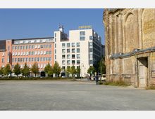 Weitläufiger Platz mit einer Ansicht eines Backsteingebäudeteils rechts im Bild, einer historischen Ruine. Links und zentral sind moderne Gebäudekomplexe mit einer Mischung aus roten und weißen Fassaden zu sehen, flankiert von einer Baumreihe entlang der Straße.