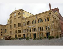 Historisches Backsteingebäude mit gelber Fassade und großen Rundbogenfenstern, beschriftet mit "PALAIS", auf einem gepflasterten Platz mit Pflanzenkübeln; im Hintergrund ein Schornstein.