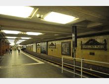 U-Bahn-Station mit gebogener Bahnsteigkante, gelblich gefliesten Wänden und einer Beschilderung mit der Aufschrift „Fehrbelliner Platz“. Die Decke ist mit rechteckigen Leuchtfeldern beleuchtet, und an den Wänden sind dekorative Details und Bilder zu sehen.