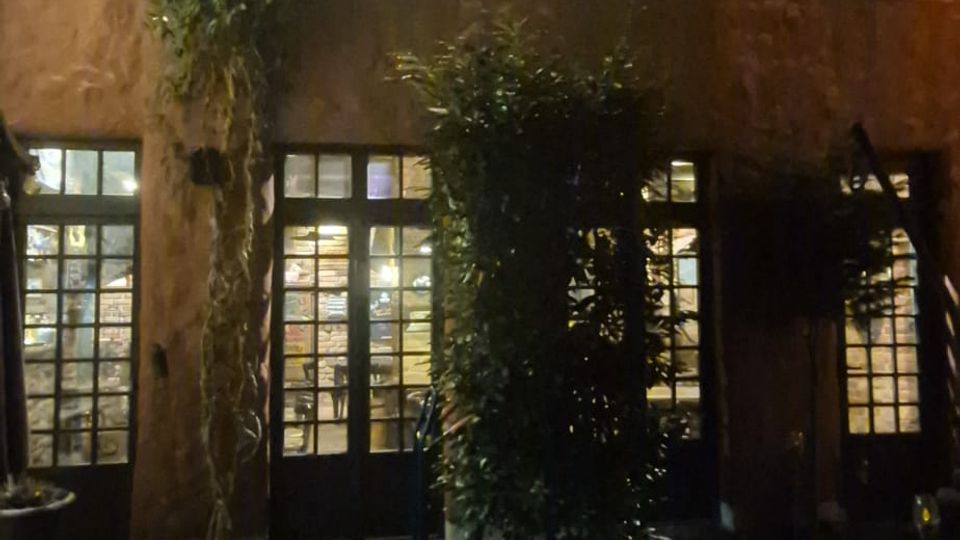 Nächtlicher Innenhof mit gepflastertem Boden vor einem zweigeschossigen Gebäude mit rau verputzter Fassade, mehreren Fenstern und großflächigen Glas-Türen im Erdgeschoss. Außenleuchten beleuchten die Wand, und Kletterpflanzen sowie große Pflanzkübel stehen entlang der Fassade.