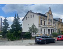 Eckhaus im Stil der Gründerzeit mit weißem und gelbem Fassadenschmuck, bodentiefen Fenstern und einem spitzen Giebel; davor stehen zwei Autos auf einer gepflasterten Straße. Rechts und links des Hauses stehen Bäume, und ein Zaun mit Tor grenzt das Grundstück ab.