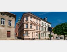 Straßenecke in einer Altstadt mit dreigeschossigen, reich verzierten Gründerzeit-Häusern in Pastellfarben, großen Fenstern und Stuckfassaden. Im Hintergrund ragt ein Backsteinturm mit grünem Kupferdach auf; am Straßenrand stehen Laterne, Verkehrszeichen und ein Baum.