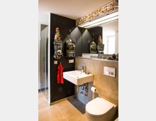 Modernes Badezimmer mit dunkler Wand, an der zwei Haken mit Rucksäcken hängen, einer roten Handtuchhalterung und einem rechteckigen Waschbecken. Ein großer Spiegel mit Fotos an der Oberkante reflektiert den Raum, während links eine Glasduschkabine und rechts eine Toilette mit Wandspülung zu sehen sind.