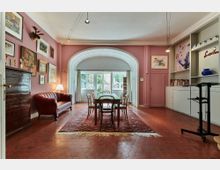 Wohnzimmer mit rosa Wänden, einem roten Sofa, einem Esstisch mit Holzstühlen auf einem orientalischen Teppich und großen Fenstern in einem bogenförmigen Durchgang. Die Einrichtung umfasst eine Wand mit Kunstwerken, eine Kommode und Regale mit Büchern und dekorativen Elementen.