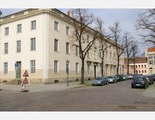 Straßenansicht mit einem historischen, hellen Gebäude im klassizistischen Stil mit hohen Fenstern und einer verzierten Dachbalustrade. Vor dem Gebäude stehen Bäume ohne Blätter, parkende Autos und Verkehrsschilder auf dem gepflasterten Straßenabschnitt.