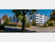 Mehrstöckige Wohngebäude mit Balkonen und einer Fassade in hellen Farben mit grauen und gelben Akzenten, umgeben von Bäumen, Sträuchern und einem Gehweg. Ein Verkehrszeichen und ein Zaun sind im Vordergrund sichtbar.