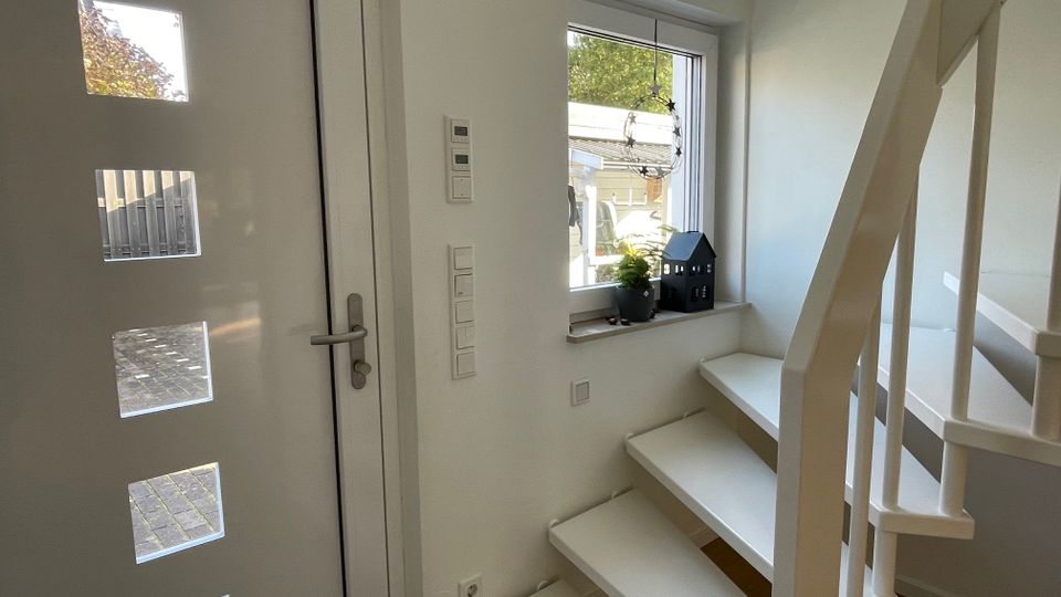 Weiß gestrichener Eingangsbereich mit moderner Haustür mit matten Glaseinsätzen, daneben ein Fenster mit Pflanzen- und Dekorationsobjekt auf der Fensterbank. Rechts davon führt eine schmale weiße Treppe nach oben.