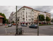Kreuzung in einem städtischen Wohngebiet mit mehrgeschossigen Altbauten im Hintergrund, darunter ein weißes Eckgebäude mit Ladengeschäften im Erdgeschoss. Verkehrsampeln, Straßenschilder und geparkte sowie fahrende Autos sind im Vordergrund zu sehen, daneben Fuß- und Radwege mit Passanten.