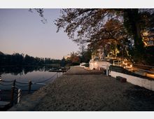 Lakeside waterfront at dusk with a sandy shoreline and calm water, bordered by small docks and rope railings. A terraced walkway with white retaining walls and wooden handrails runs along a tree-covered slope with warm lights and buildings set back above the shore.