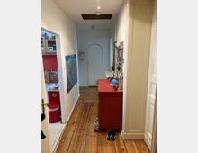 Flur in einer Wohnung mit Holzdielenboden, links ein Durchgang zur Küche mit roten Elementen, rechts ein rotes Sideboard mit darauf abgelegten Gegenständen. An der Wand hängt eine Weltkarte, im Hintergrund ist eine weiße Tür mit einem Rundbogen sichtbar.