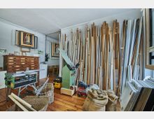 Ein Holzwerkstattraum mit gestapelten Holzleisten an der Wand, verschiedenen Maschinen wie einer Kreissäge und Hobelmaschine, geflochtenen Körben und Jutesäcken auf einem Holzboden. An der Wand hängen Bilderrahmen und ein hölzernes Schubladensystem steht im Hintergrund.