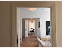 Blick durch eine Türöffnung in eine Wohnungsinnenansicht mit Holzboden, weißen Wänden und hohen Decken. Im Vordergrund ein beiges Sofa, im Hintergrund ein Essbereich mit einem Tisch, Stühlen und einem Kronleuchter.