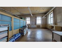 Geräumiges, vernachlässigtes Büro mit hohen Decken, großen Fenstern mit Vorhängen und freiliegenden Heizkörpern an den Wänden; links ein Raumteiler mit Glasflächen und gestapelten Akten, rechts mehrere Kartons und ein leerer Tisch.