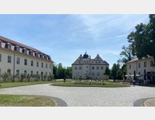 Großer gepflasterter Hof mit einem historischen Gebäude in der Mitte, das ein Mansarddach und eine auffällige Kuppel hat. Links und rechts umrahmen zwei ähnliche Gebäude mit weißen Fassaden und roten Ziegeldächern den Platz; rechts gibt es zusätzlich eine Außengastronomie.