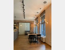 Ein modernes Café mit Holzboden, freiliegenden Backsteinwänden und großen Fenstern, die Tageslicht hereinlassen. Mehrere Tische mit schwarzen Stühlen stehen im Raum, zusammen mit hängenden Glühbirnenlampen an der Decke.