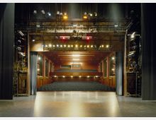 Blick von der Bühne in einen Theatersaal mit leeren Sitzreihen, Balkon und dunklen Holzvertäfelungen. Die Decke ist mit Bühnentechnik, Beleuchtung und Scheinwerfern ausgestattet.