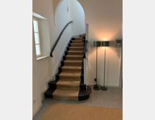 Treppe in einem Innenraum mit dunkel lackierten Stufen und einem beigefarbenen Teppichläufer, der von Metallstangen gehalten wird. Links befindet sich ein Fenster mit Sprossen, rechts stehen zwei Stehlampen neben einer Gegensprechanlage.