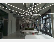 Ein modernes, offenes Loft mit Kleiderständern entlang der Fensterfront, das als Showroom oder Büro dient. Die Decke zeigt freiliegende Rohre und asymmetrische Leuchtstäbe, während der Raum durch große Fenster gut beleuchtet ist.