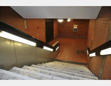 Treppenabgang in eine U-Bahn- oder Unterführung mit roten Ziegelwänden, beleuchtet durch Deckenlampen und Handläufe mit integrierten Leuchten. Der Boden ist mit roten Fliesen ausgelegt, und an der Wand hängt ein schwarzes Informationsschild.