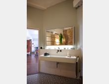 Modernes Badezimmer mit einem rechteckigen, weißen Waschbecken; darüber ein großer Spiegel, der einen Backstein-Wandabschnitt im Raum reflektiert. Der Boden ist mit gemusterten Fliesen gestaltet, und im Spiegel hängt eine grüne Pflanze. Im Hintergrund öffnet sich der Raum zu einem Bereich mit Holzboden.