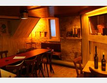 Gemütlicher Raum mit rustikalem Design, geprägt von Backsteinwänden, Holzmöbeln und einer langen, rot abgedeckten Tischreihe. Die warme Beleuchtung und dekorative Elemente wie Flaschen und Wandbilder verleihen dem Raum eine einladende Atmosphäre.