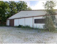 Ein einstöckiges, weiß gestrichenes Industriegebäude mit flachem Satteldach, großen Holztoren und einem Fenster mit Metallrahmen. Der Vorplatz besteht aus Pflastersteinen, teils überwuchert von Gras und Unkraut, und ist umgeben von Bäumen.