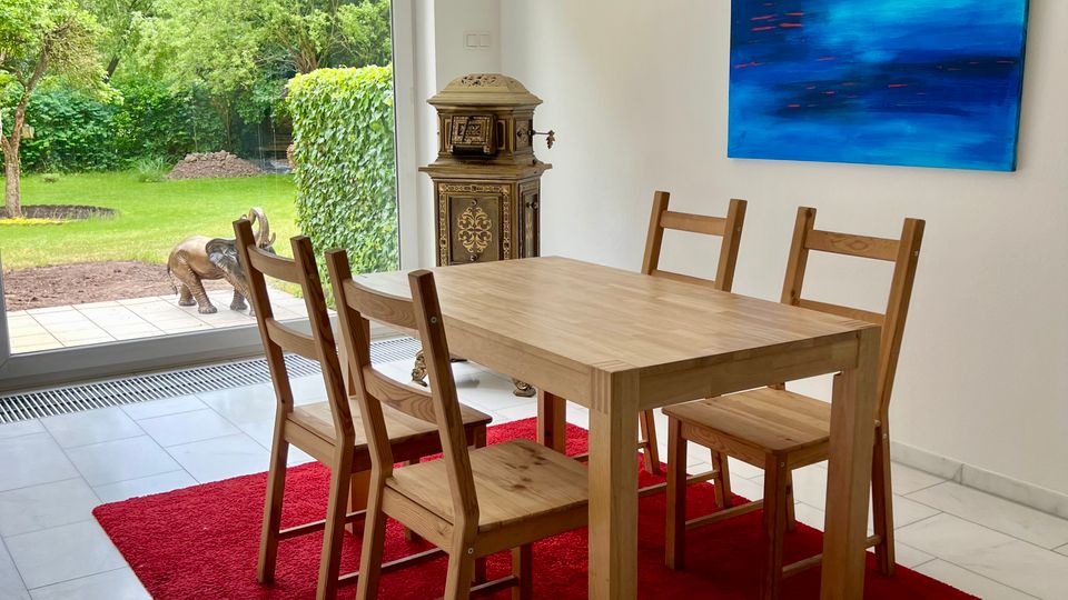 Heller Essbereich mit Holztisch und vier Holzstühlen auf einem roten Teppich, weißen Wänden und Fliesenboden. Große Schiebetür öffnet den Blick in einen grünen Garten; an der Wand hängt ein blaues abstraktes Bild, daneben steht ein antiker Ofen.