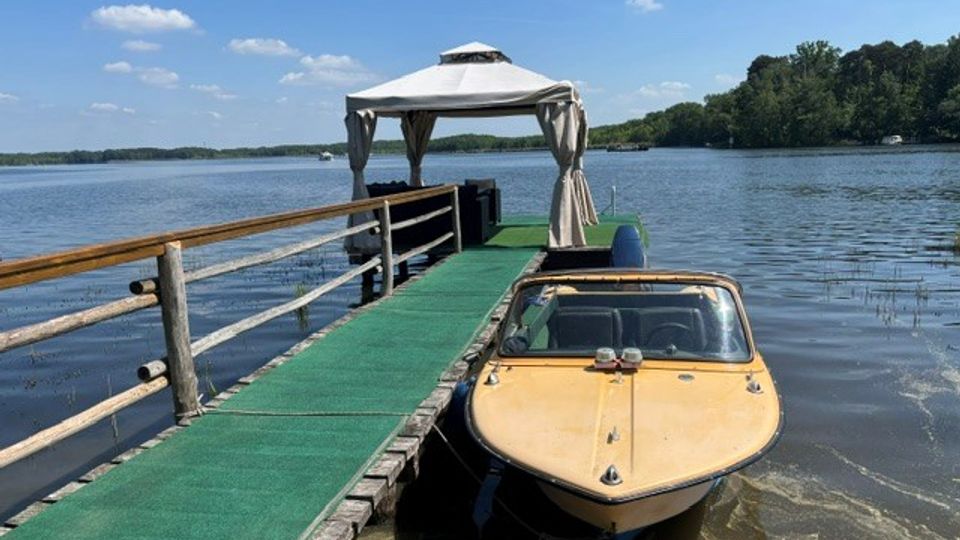 Holzsteg mit grünem Bodenbelag führt über einen ruhigen See zu einem kleinen Pavillon mit weißem Dach und Vorhängen. Rechts liegt ein Motorboot am Steg, im Hintergrund sind bewaldete Ufer und blauer Himmel zu sehen.