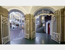 Ein historischer Innenraum mit geschwungenen weißen Schmiedeeisen-Toren und Treppen. Im Hintergrund sind steinerne Säulen, verzierte Geländer und runde Türen sichtbar, die den Raum architektonisch definieren.