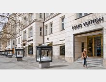 Straßenansicht eines Luxusgeschäfts der Marke Louis Vuitton, in einem Gebäude mit heller Steinfassade und großen Schaufenstern. Vor dem Geschäft stehen gläserne Ausstellungsvitrinen, und ein Fußgänger passiert den Eingang.